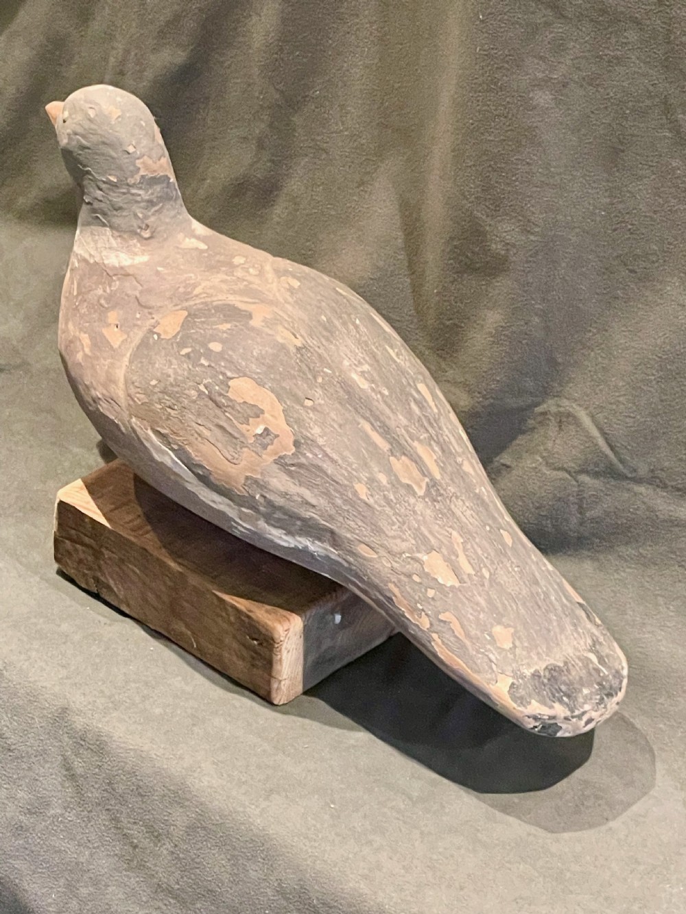 A hand-carved wooden pigeon decoy featuring weathered grey paint, mounted on a small rectangular wooden block. View IX