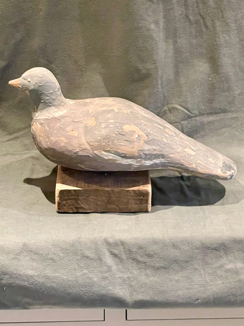 A hand-carved wooden pigeon decoy featuring weathered grey paint, mounted on a small rectangular wooden block. View X