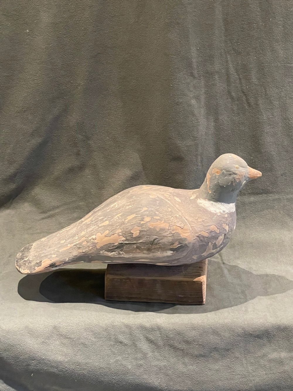 A hand-carved wooden pigeon decoy featuring weathered grey paint, mounted on a small rectangular wooden block. View XI
