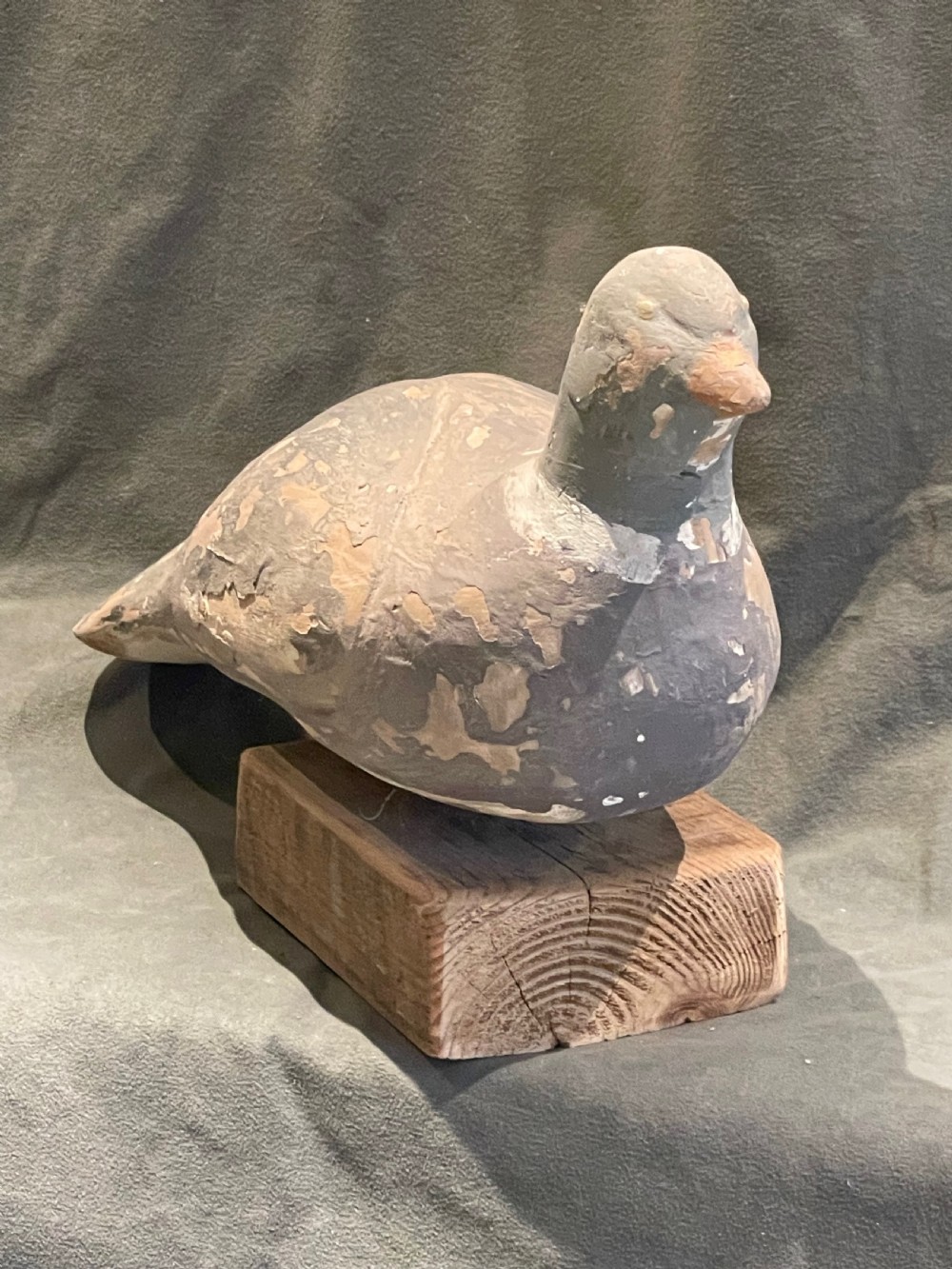A hand-carved wooden pigeon decoy featuring weathered grey paint, mounted on a small rectangular wooden block. View XII