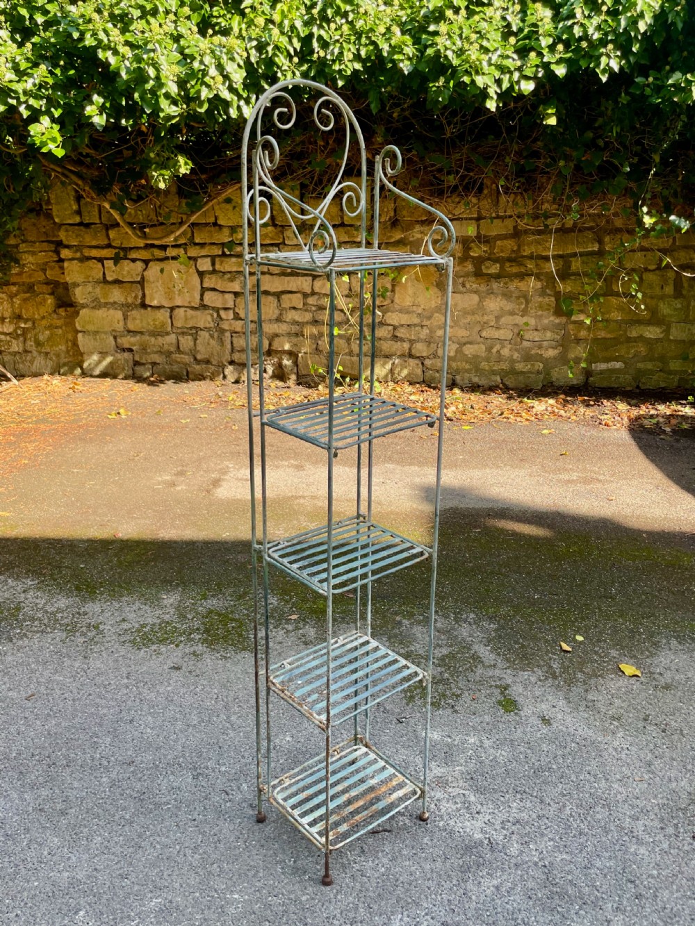 A tall, narrow painted metal plant stand with five slatted shelves and scrollwork decorative detailing at the top.