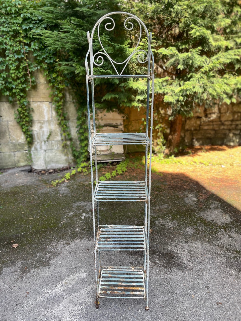 A tall, narrow painted metal plant stand with five slatted shelves and scrollwork decorative detailing at the top. View V