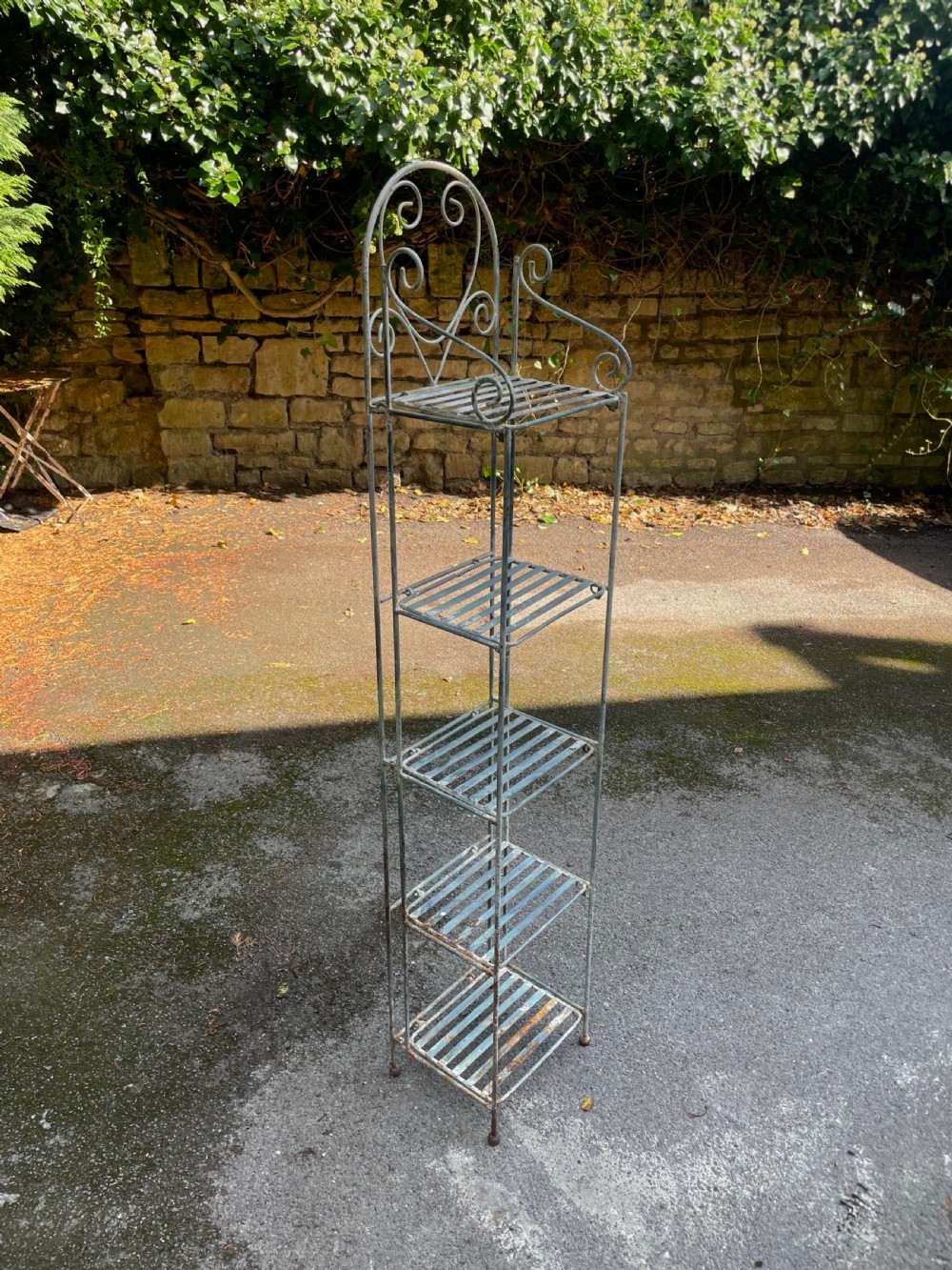 A tall, narrow painted metal plant stand with five slatted shelves and scrollwork decorative detailing at the top. View VI