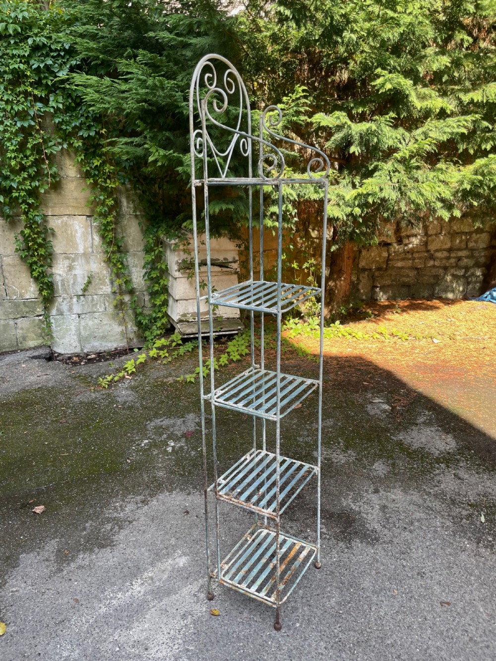A tall, narrow painted metal plant stand with five slatted shelves and scrollwork decorative detailing at the top. View VII