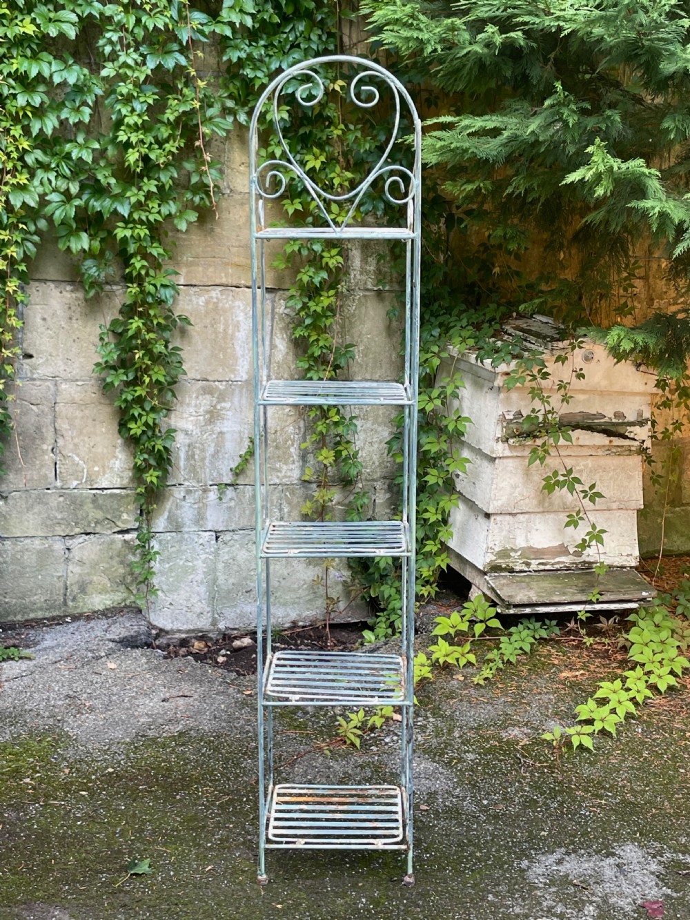 A tall, narrow painted metal plant stand with five slatted shelves and scrollwork decorative detailing at the top. View IX