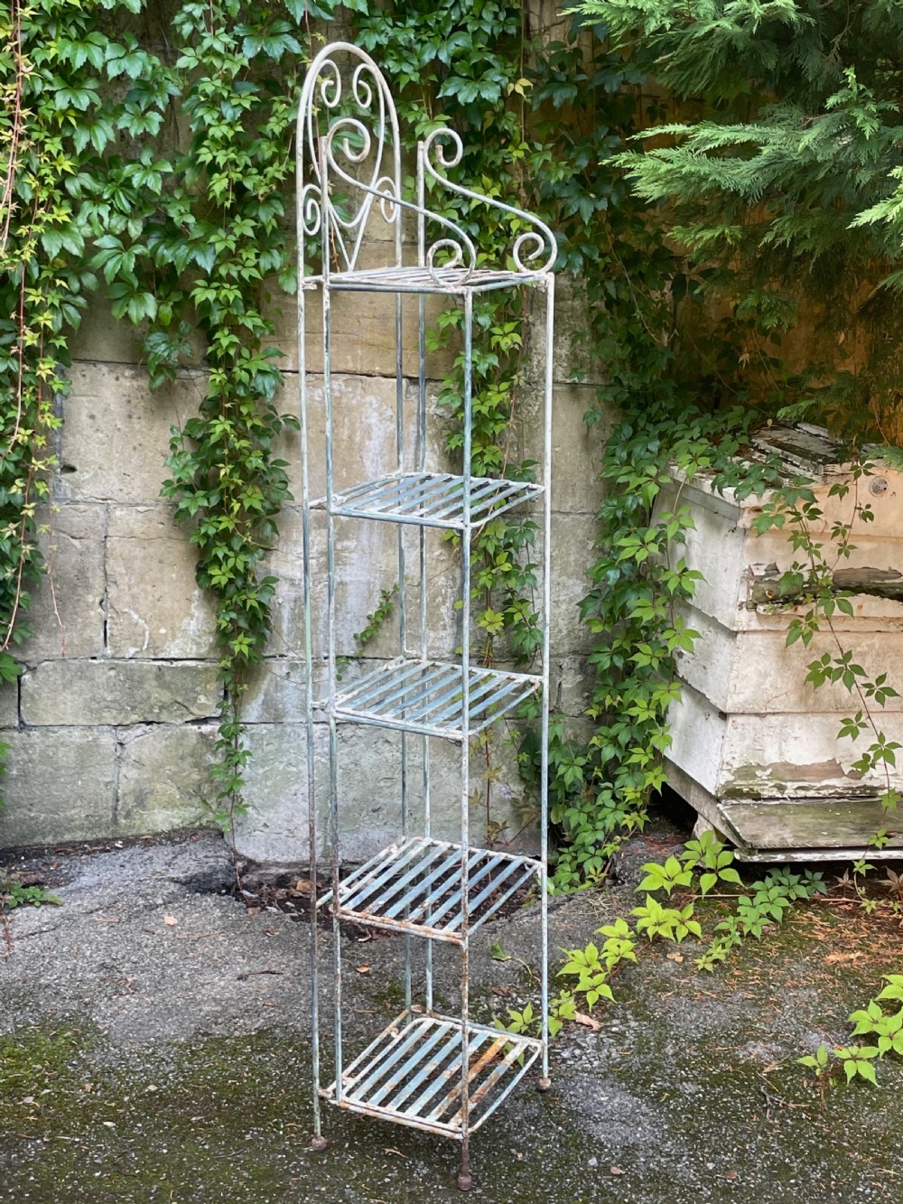A tall, narrow painted metal plant stand with five slatted shelves and scrollwork decorative detailing at the top. View X