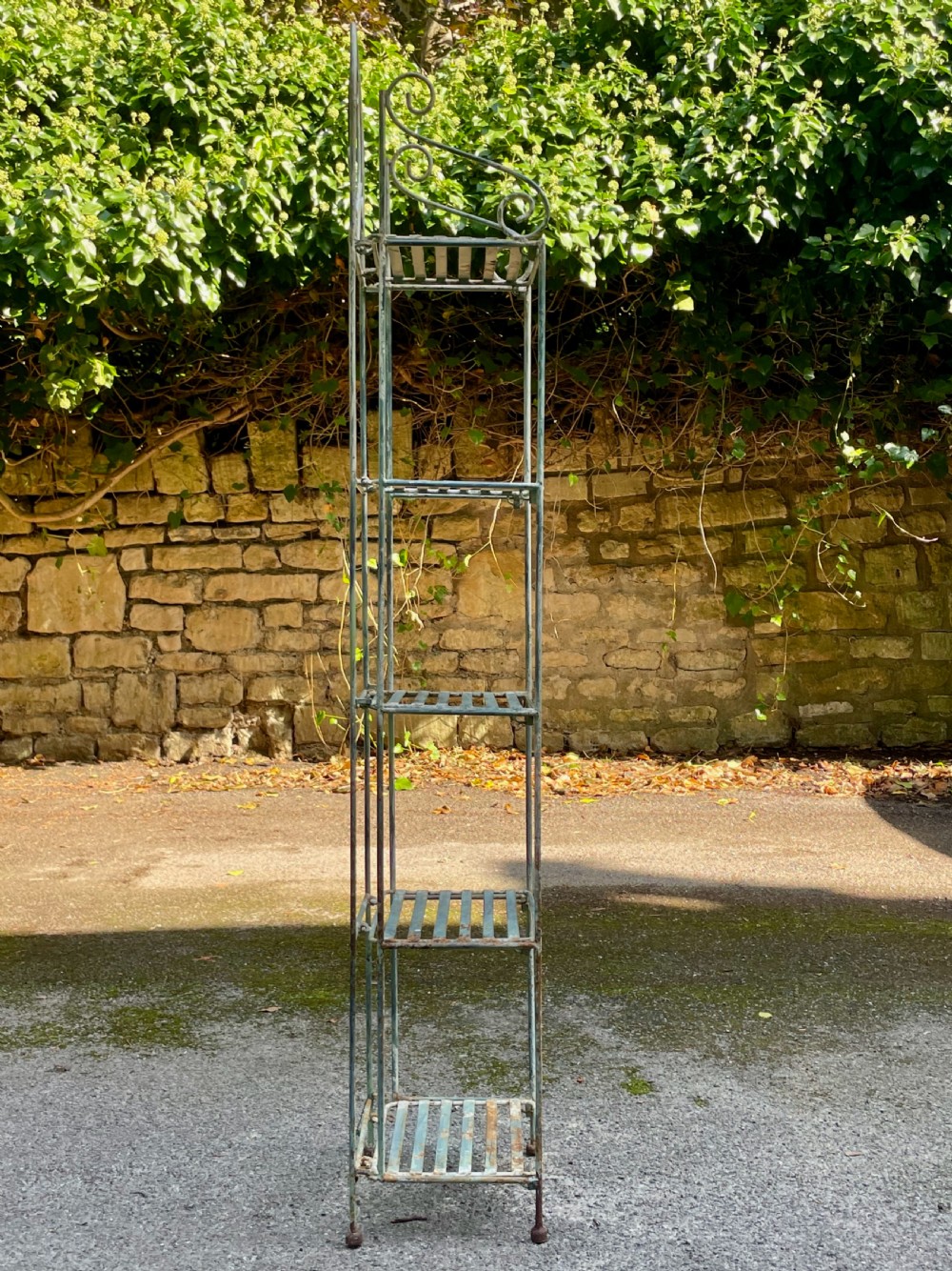 A tall, narrow painted metal plant stand with five slatted shelves and scrollwork decorative detailing at the top. View XIV