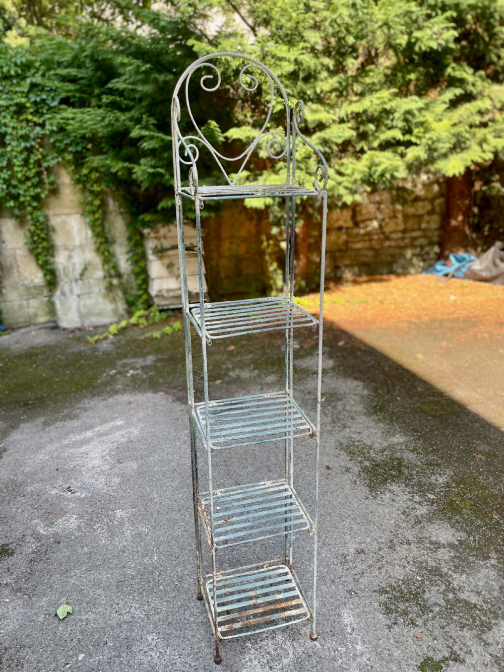 A tall, narrow painted metal plant stand with five slatted shelves and scrollwork decorative detailing at the top. View XVI
