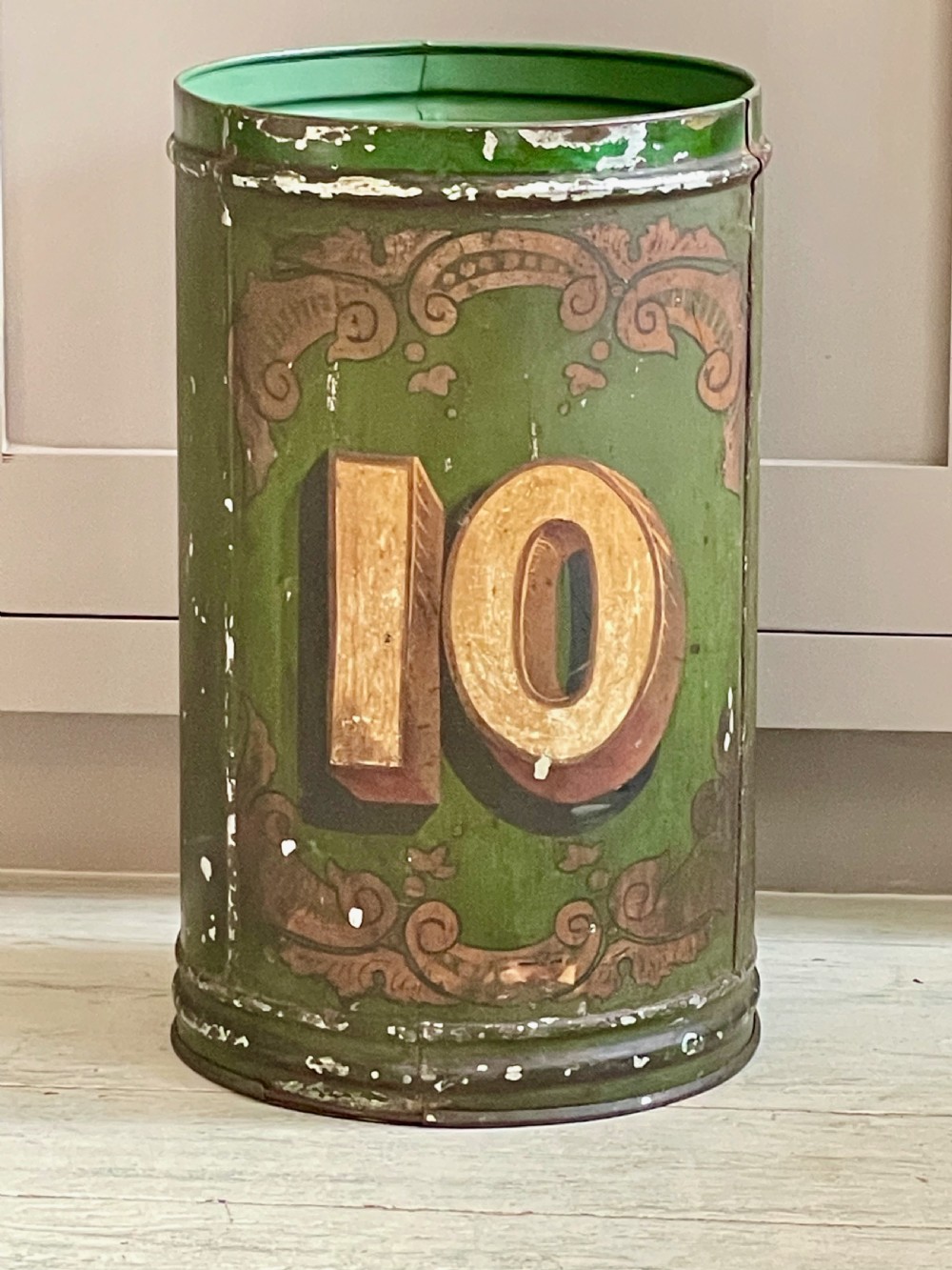 A cylindrical green painted antique metal tea canister featuring gold scrollwork and the painted number ten.