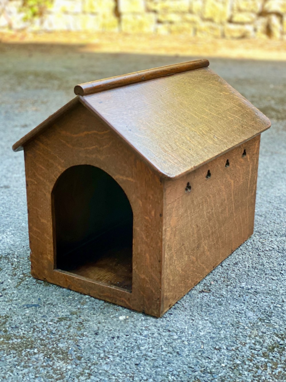 An antique Victorian warm brown oak dog kennel featuring a pitched roof, arched entrance aperture, and trefoil ventilation cutouts along the side.