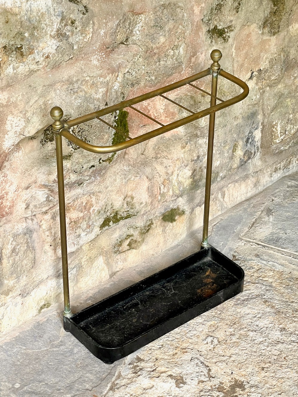 An antique Edwardian brass stick stand featuring a divided top rail with urn finials and a black cast iron drip tray base.