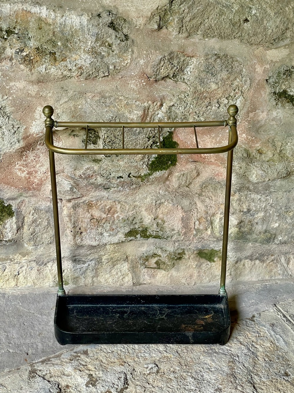 An antique Edwardian brass stick stand featuring a divided top rail with urn finials and a black cast iron drip tray base. View II