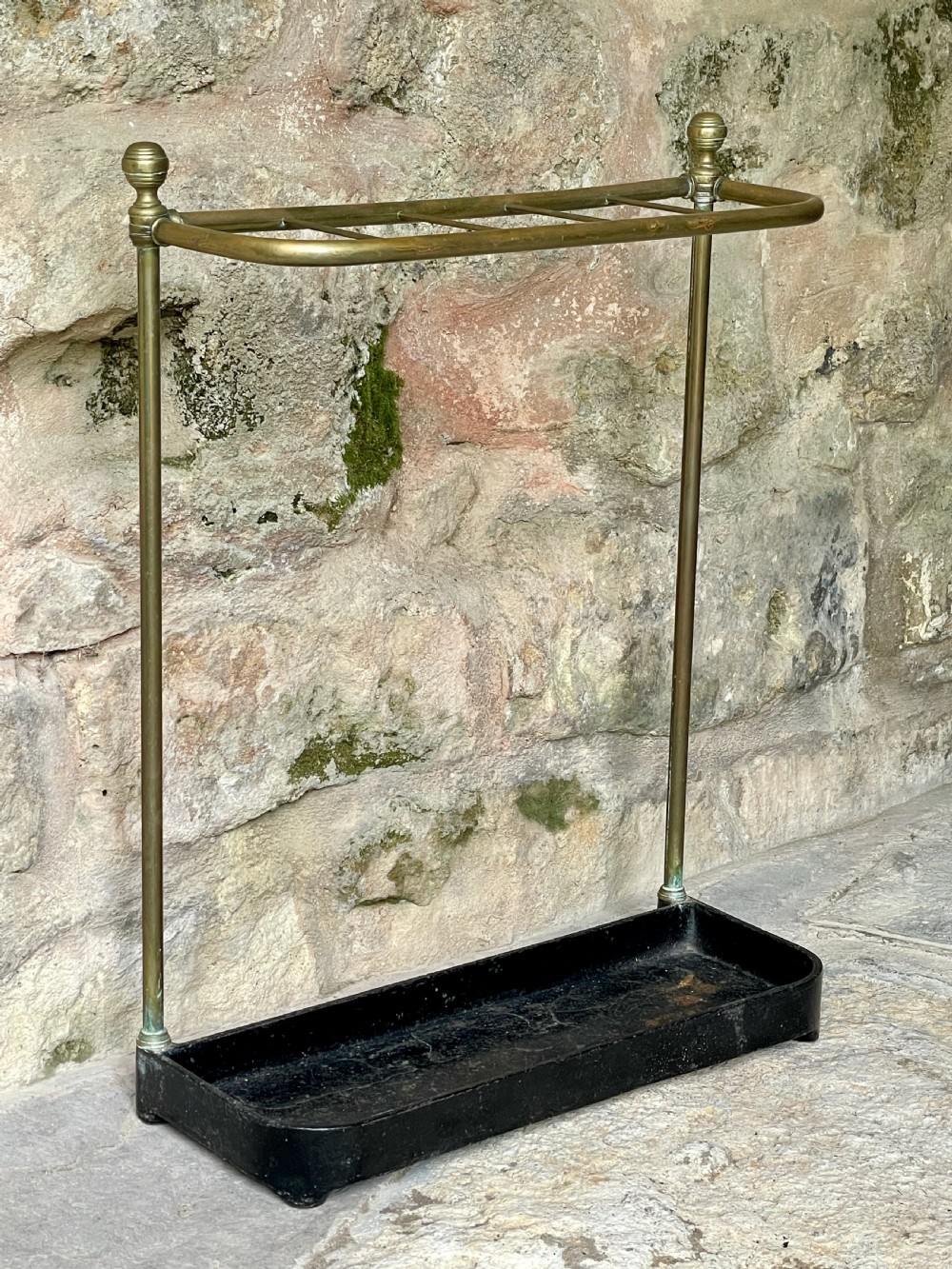 An antique Edwardian brass stick stand featuring a divided top rail with urn finials and a black cast iron drip tray base. View IV