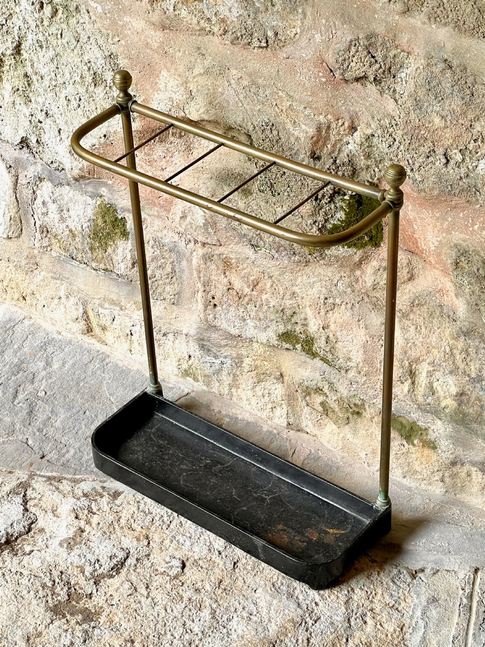 An antique Edwardian brass stick stand featuring a divided top rail with urn finials and a black cast iron drip tray base. View V
