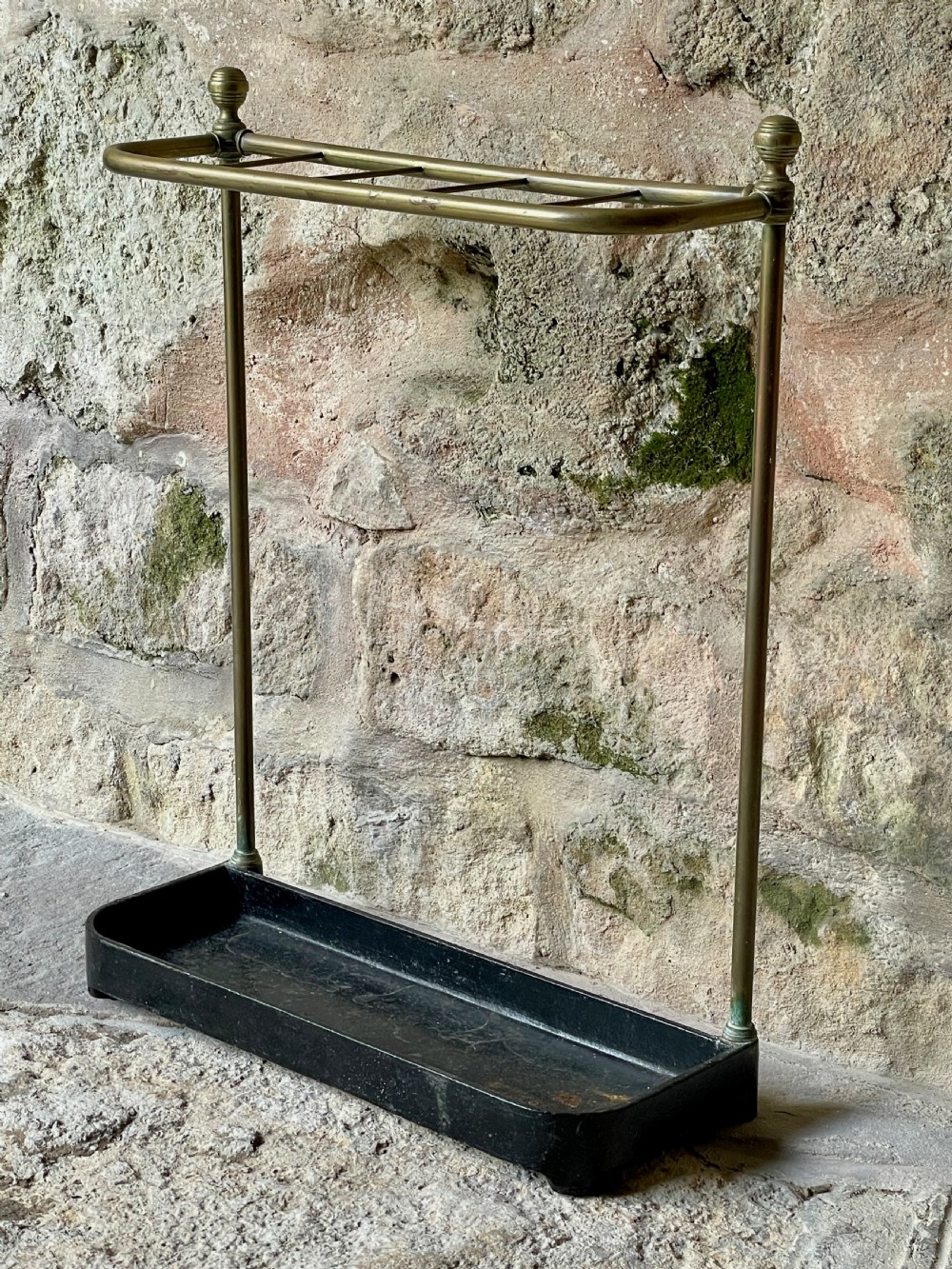 An antique Edwardian brass stick stand featuring a divided top rail with urn finials and a black cast iron drip tray base. View VI