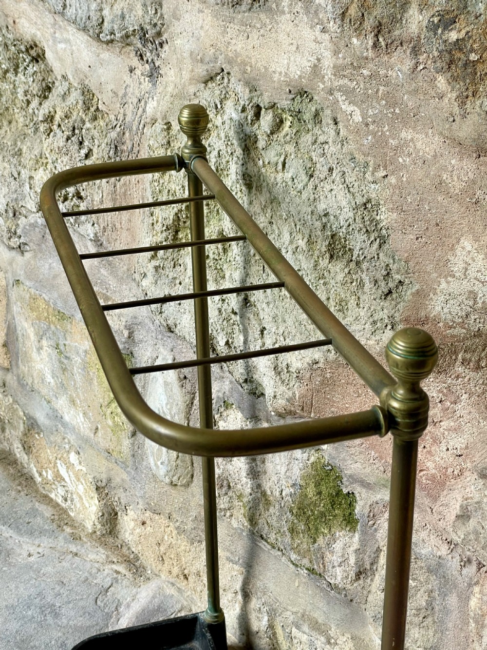 An antique Edwardian brass stick stand featuring a divided top rail with urn finials and a black cast iron drip tray base. View IX