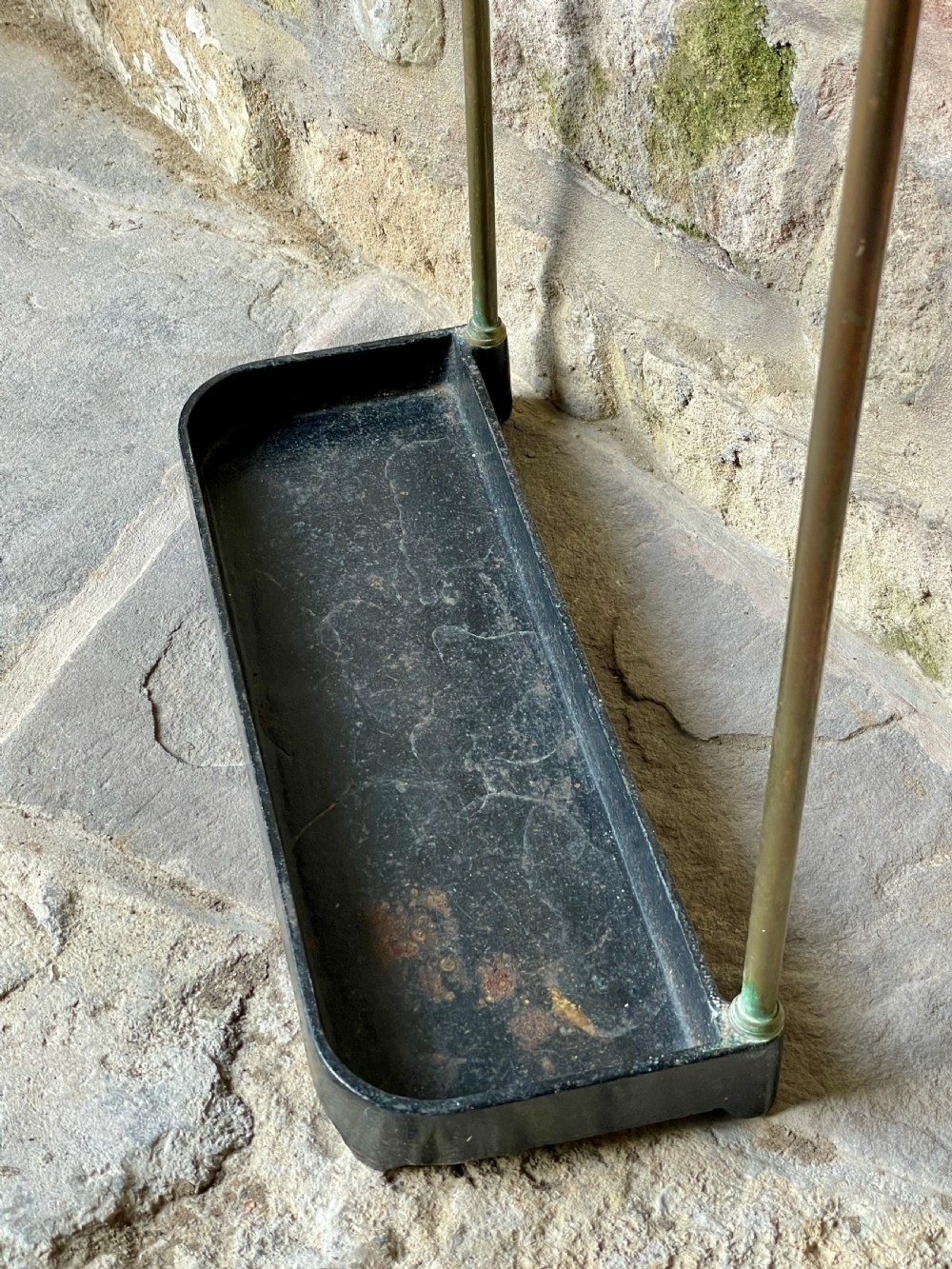 An antique Edwardian brass stick stand featuring a divided top rail with urn finials and a black cast iron drip tray base. View X