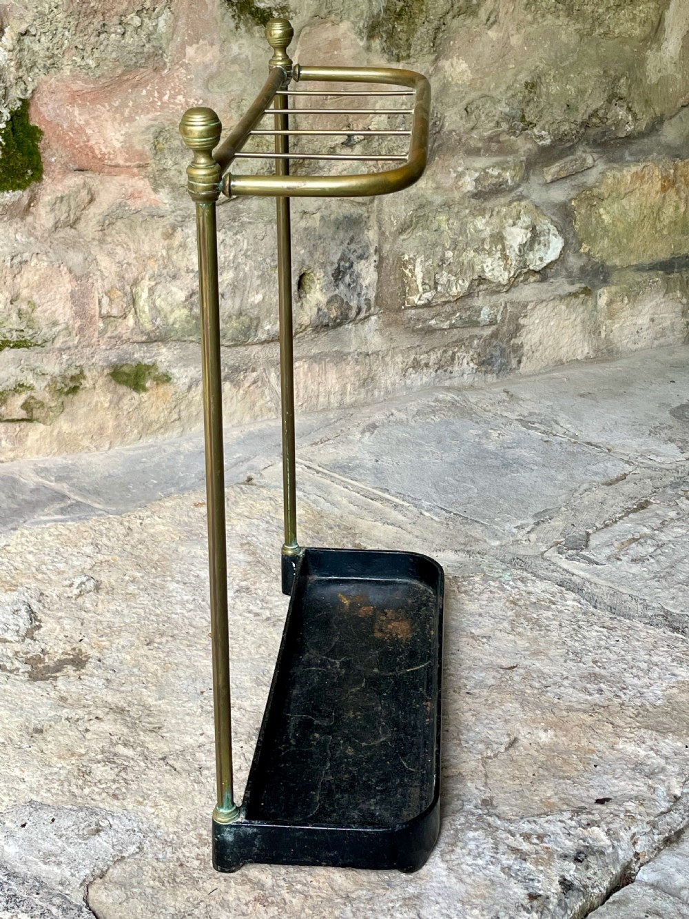 An antique Edwardian brass stick stand featuring a divided top rail with urn finials and a black cast iron drip tray base. View XI