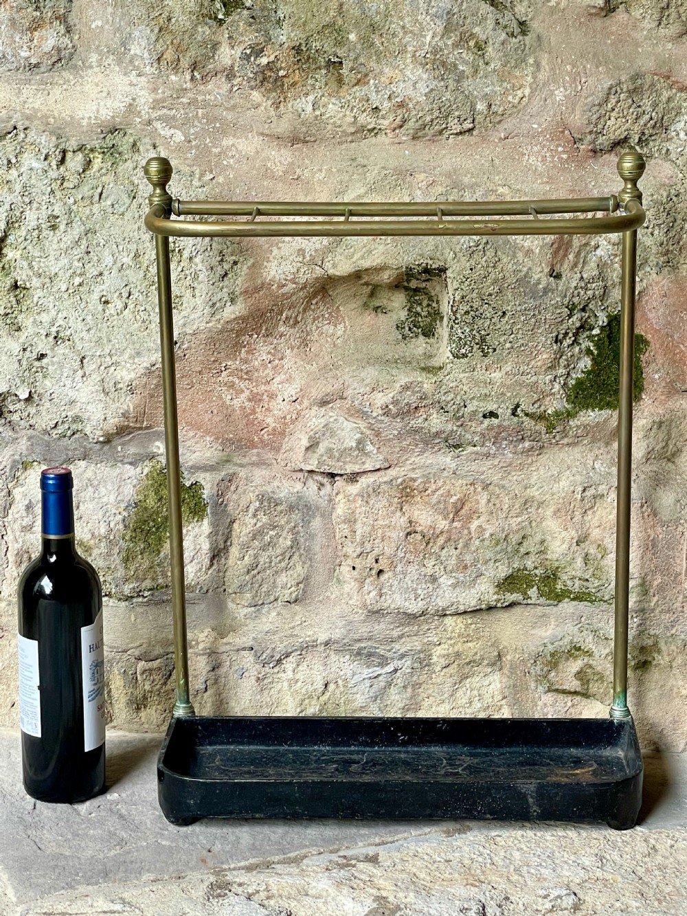 An antique Edwardian brass stick stand featuring a divided top rail with urn finials and a black cast iron drip tray base. View XV