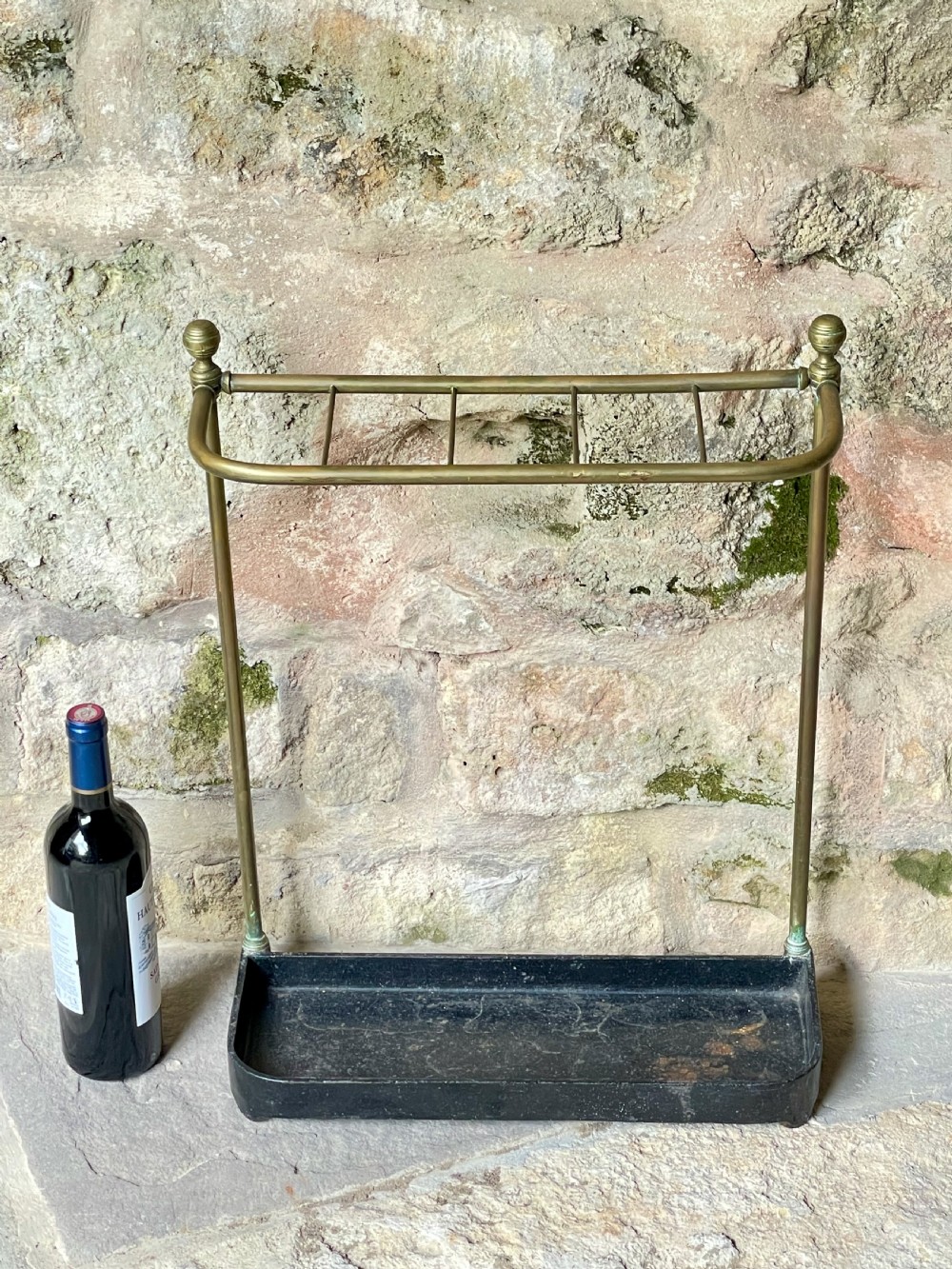 An antique Edwardian brass stick stand featuring a divided top rail with urn finials and a black cast iron drip tray base. View XVI