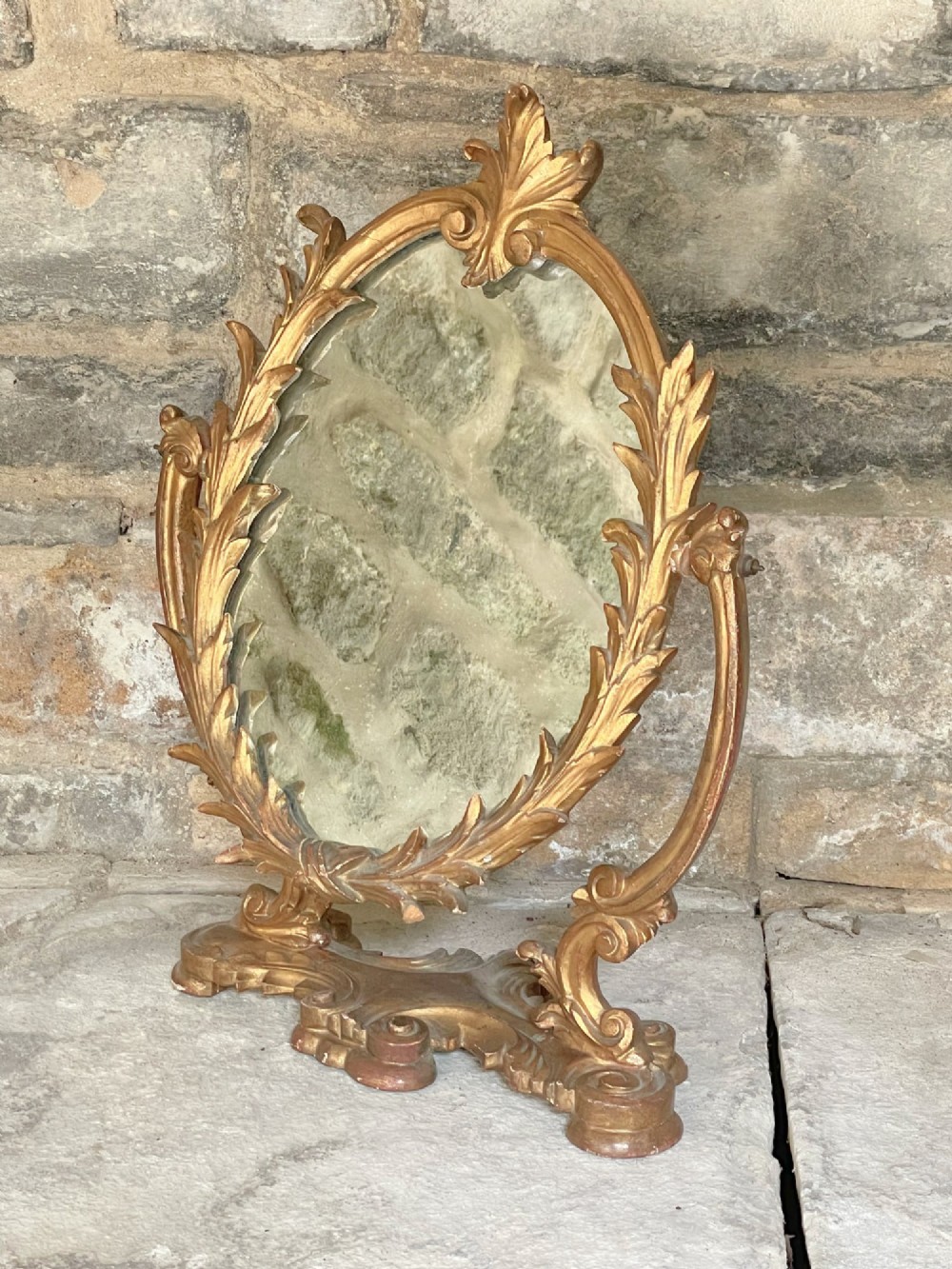 A carved giltwood toilet mirror with an oval pivoting mirror frame set on a decorative scrolling base.
