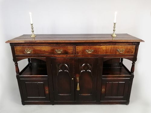 fig 3. A George III dark oak low dresser featuring central cupboard doors with Gothic pointed arch panels, upper drawers with brass swan-neck handles, and turned supports.