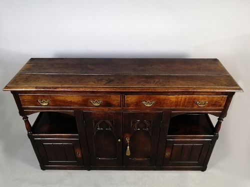 fig 5. A George III dark oak low dresser featuring central cupboard doors with Gothic pointed arch panels, upper drawers with brass swan-neck handles, and turned supports.
