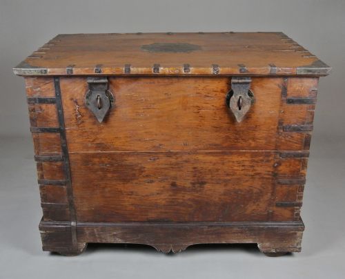 fig 1. A large brown teak wood chest reinforced with dark iron straps, corner brackets, side handles, and front latches.