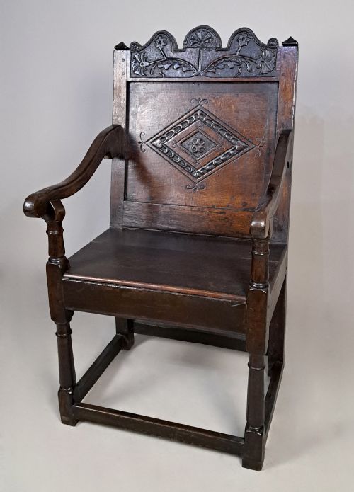 fig 2. A dark oak wainscot armchair featuring a carved lozenge motif on the back panel, shaped cresting rail, shaped armrests, and turned front legs with lower stretchers.