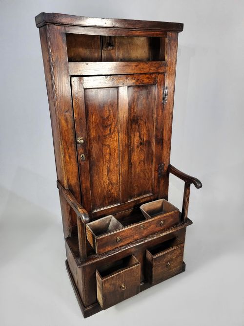 fig 1. A tall George II solid elm bacon settle featuring a high panelled cupboard back, shaped open armrests, and two integrated drawers in the base.