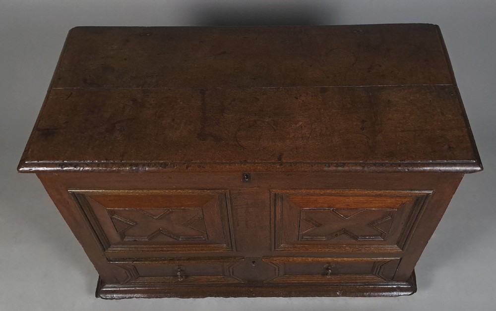 A 17th-century dark oak mule chest featuring geometric carved panel fronts, a lower drawer with brass drop handles, and a flat hinged top. View IV