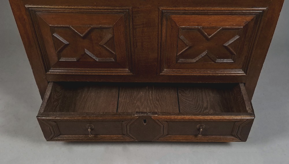 A 17th-century dark oak mule chest featuring geometric carved panel fronts, a lower drawer with brass drop handles, and a flat hinged top. View VIII