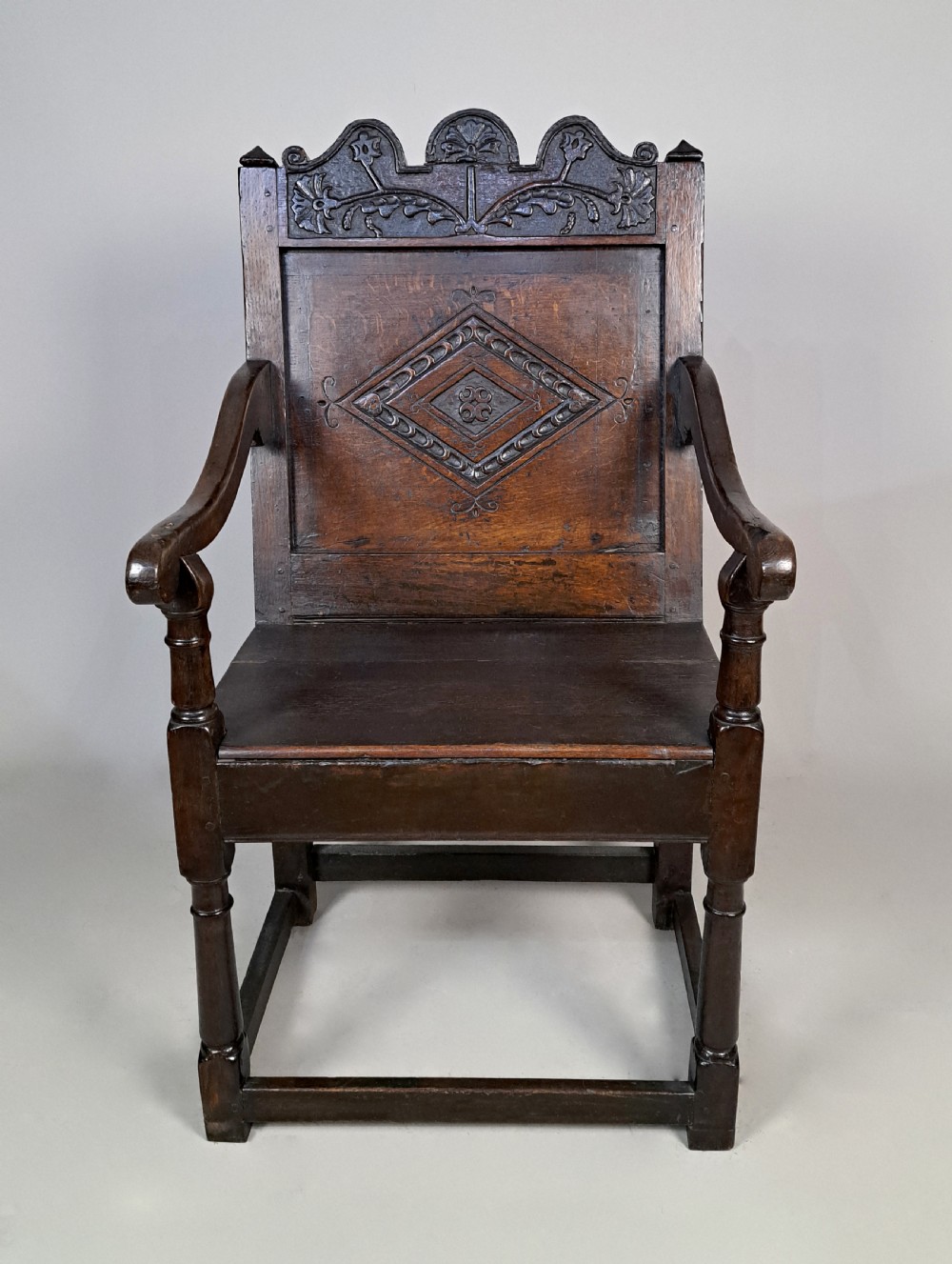 A 17th-century dark oak wainscot armchair featuring a carved lozenge back panel, shaped cresting rail, curved armrests, and turned legs. View III
