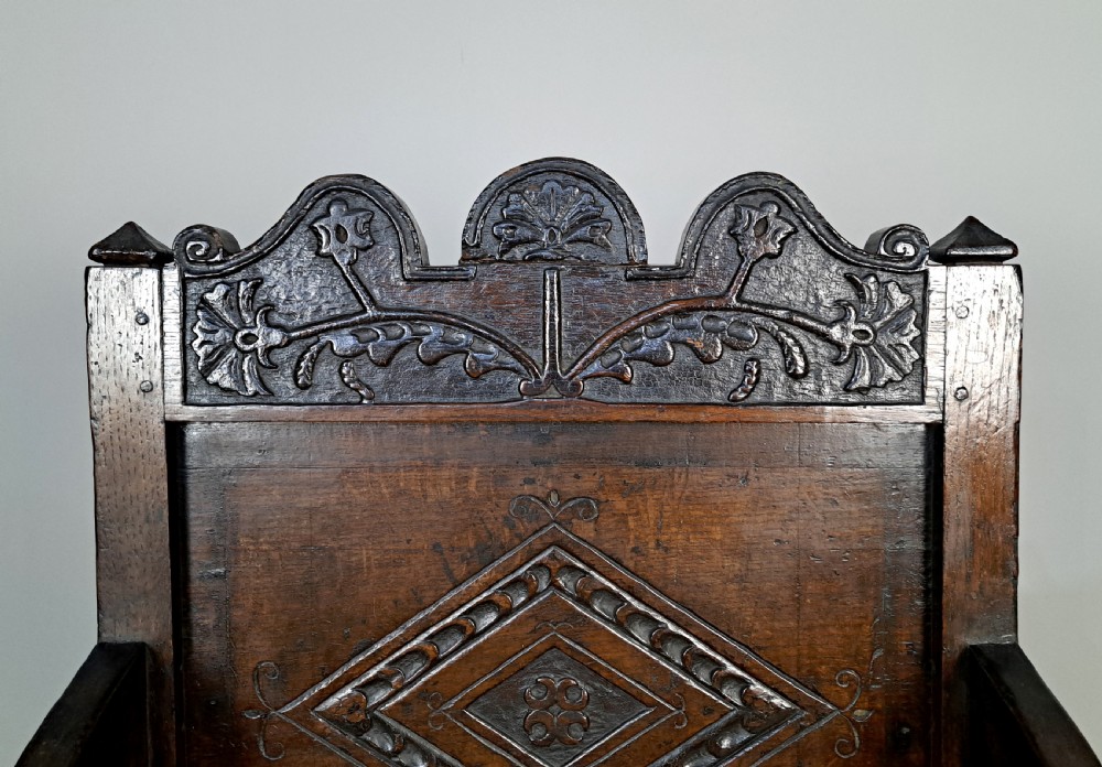 A 17th-century dark oak wainscot armchair featuring a carved lozenge back panel, shaped cresting rail, curved armrests, and turned legs. View V