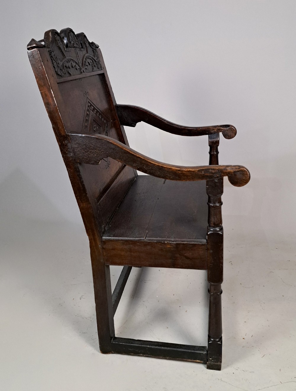 A 17th-century dark oak wainscot armchair featuring a carved lozenge back panel, shaped cresting rail, curved armrests, and turned legs. View VI
