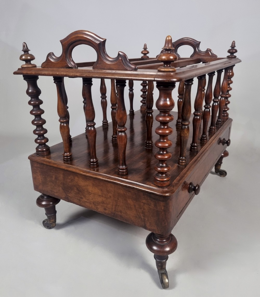 A Victorian walnut music Canterbury featuring turned spindles, division compartments, a lower drawer with twin knobs, and turned legs with brass castors. View V