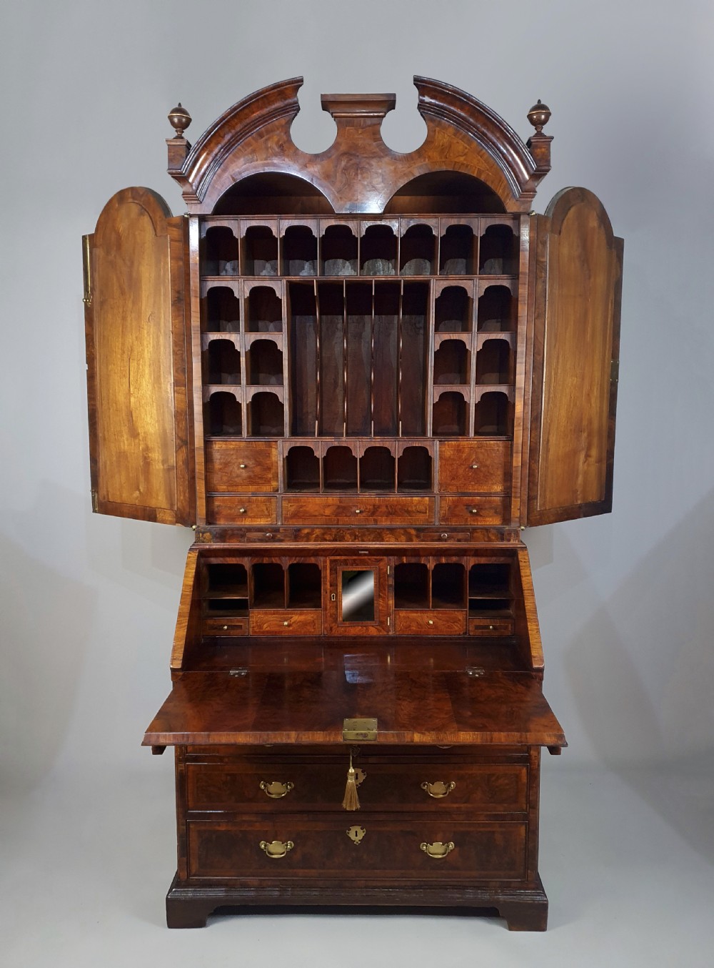 An early 18th-century Queen Anne burr walnut bureau cabinet featuring a double-domed pediment, open doors revealing multiple pigeonholes, a sloped drop-front writing desk, and lower storage drawers.