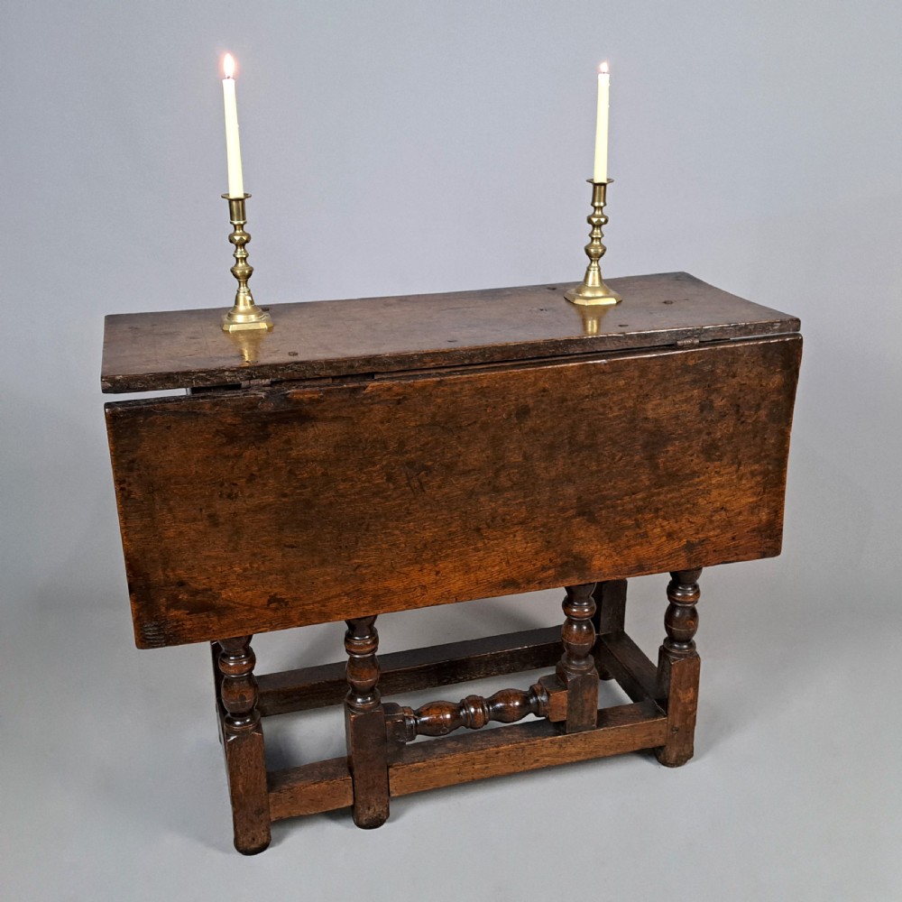 A 17th-century Charles II dark oak drop-leaf side table featuring turned legs, a hinged top flap, and sturdy lower stretchers.