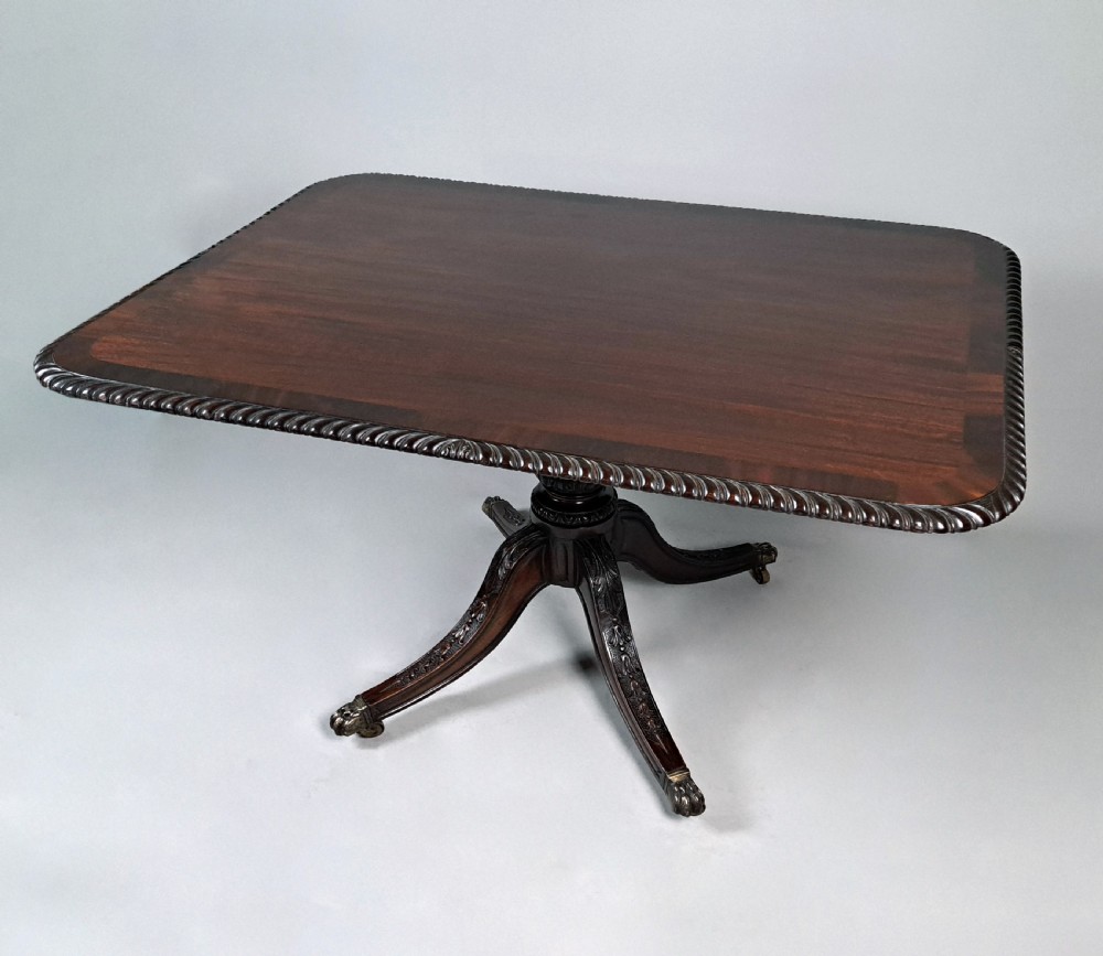 A George III mahogany dining table featuring a rectangular top with carved rope-twist edging, resting on a turned central column and four carved legs with brass castors.