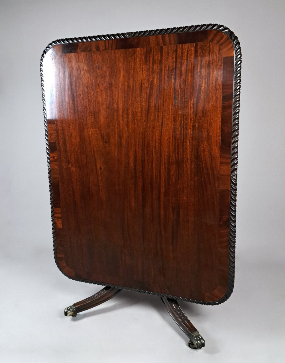 A George III mahogany dining table featuring a rectangular top with carved rope-twist edging, resting on a turned central column and four carved legs with brass castors. View V