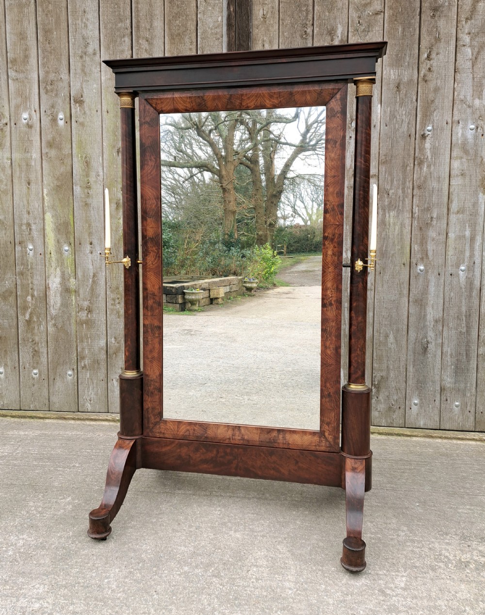 A full-length mahogany cheval mirror featuring fluted columns with brass mounts, twin brass candle sconces, and an architectural top cornice.