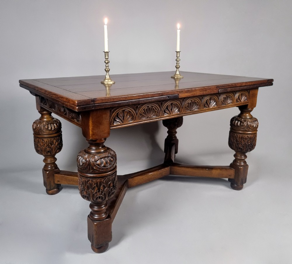 A carved warm brown oak extending dining table featuring heavy bulbous cup-and-cover legs, carved frieze detailing, and a floor stretcher base.