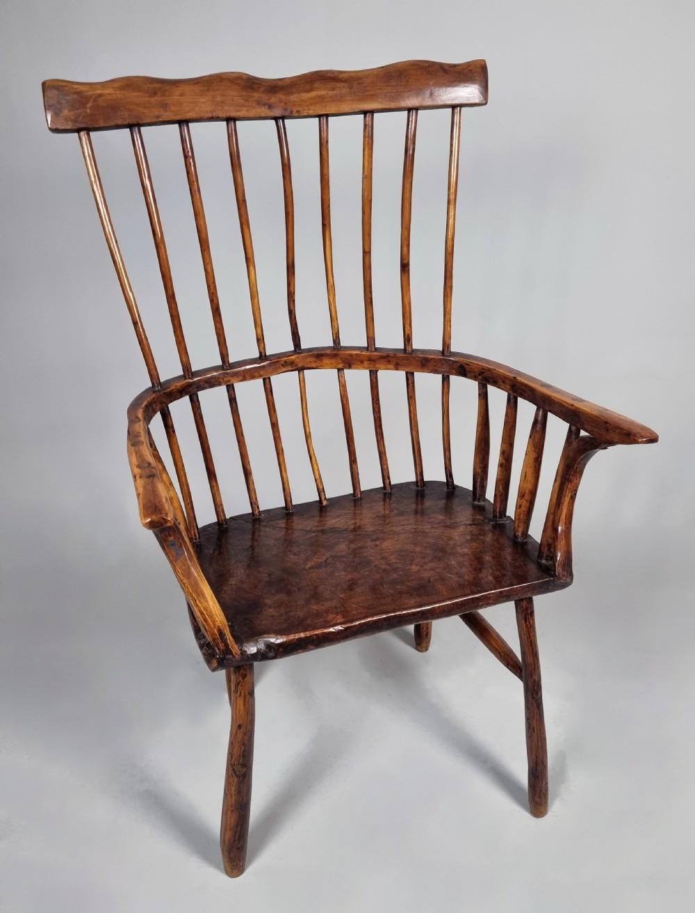 A primitive 18th-century Welsh stick armchair made from dark polished wood featuring a comb back, tapered stick spindles, and an organic curved frame.