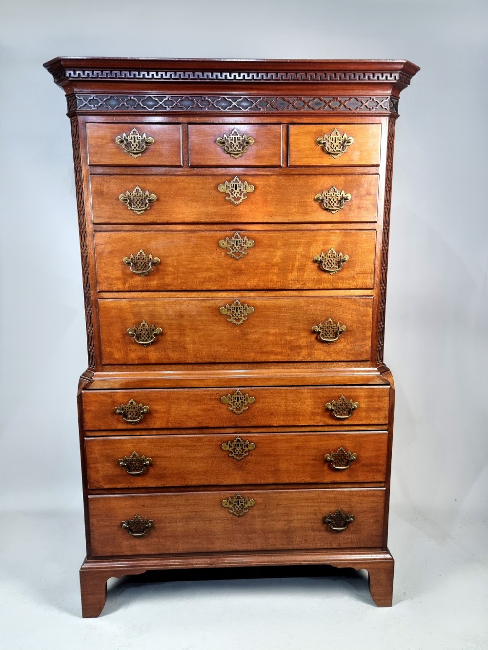 A tall George III mahogany chest on chest featuring stacked drawer sections, pierced brass handles, carved fretwork at the frieze, and bracket feet. View V