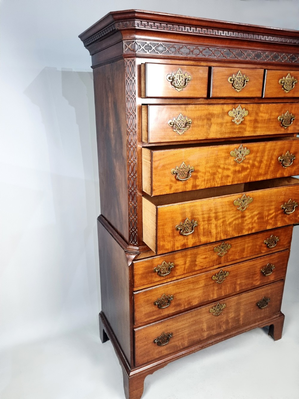 A tall George III mahogany chest on chest featuring stacked drawer sections, pierced brass handles, carved fretwork at the frieze, and bracket feet. View VII