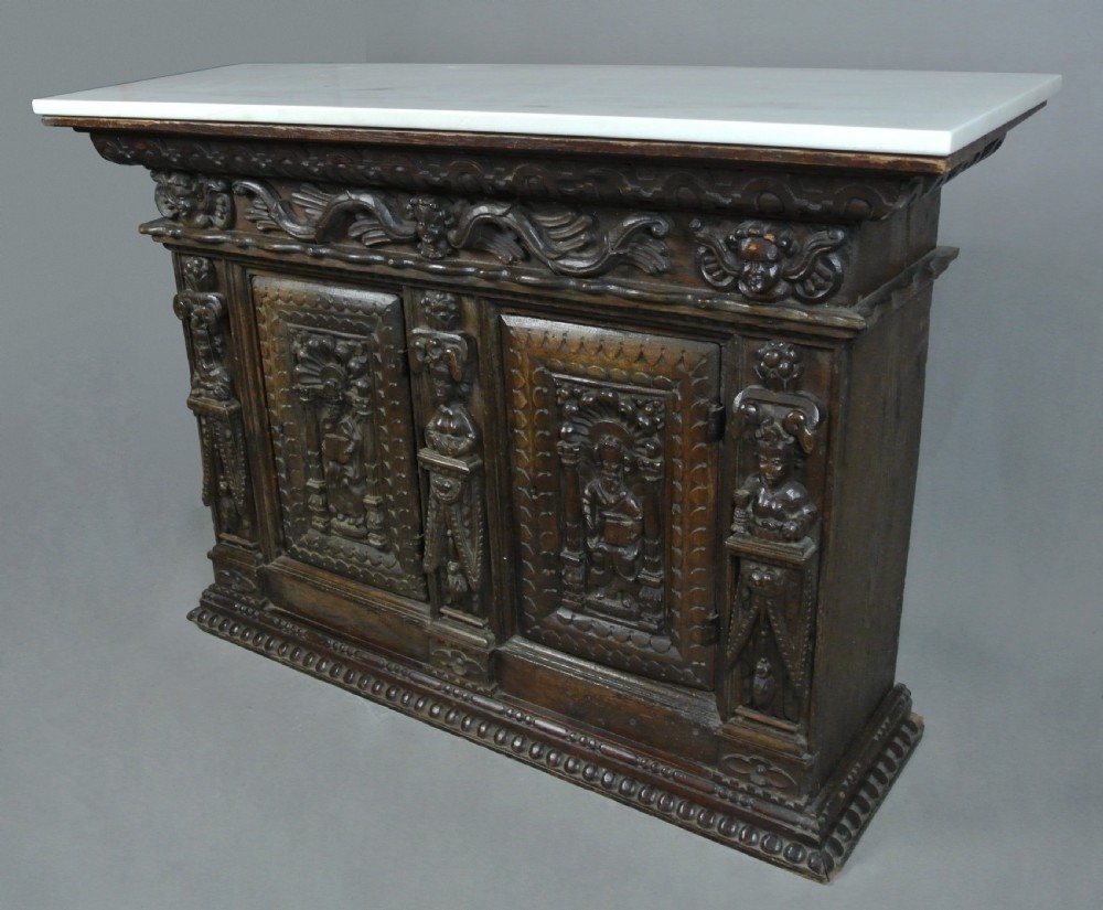 A dark carved oak 17th-century style livery cupboard featuring figural relief carvings on the panel doors and caryatids, topped with a smooth white marble slab. View VII