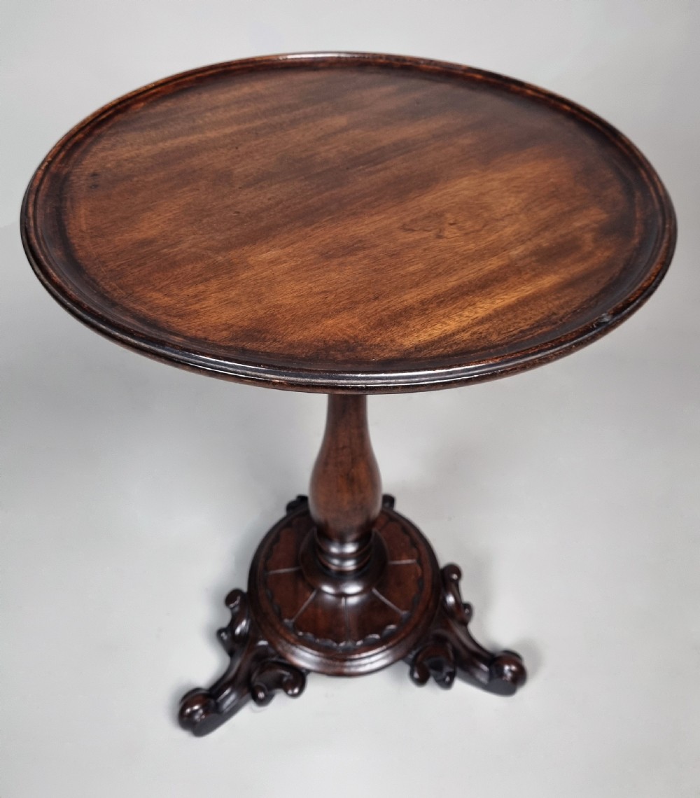A William IV mahogany wine table featuring a dished circular top, turned central pedestal column, and a carved triform base with scroll feet.