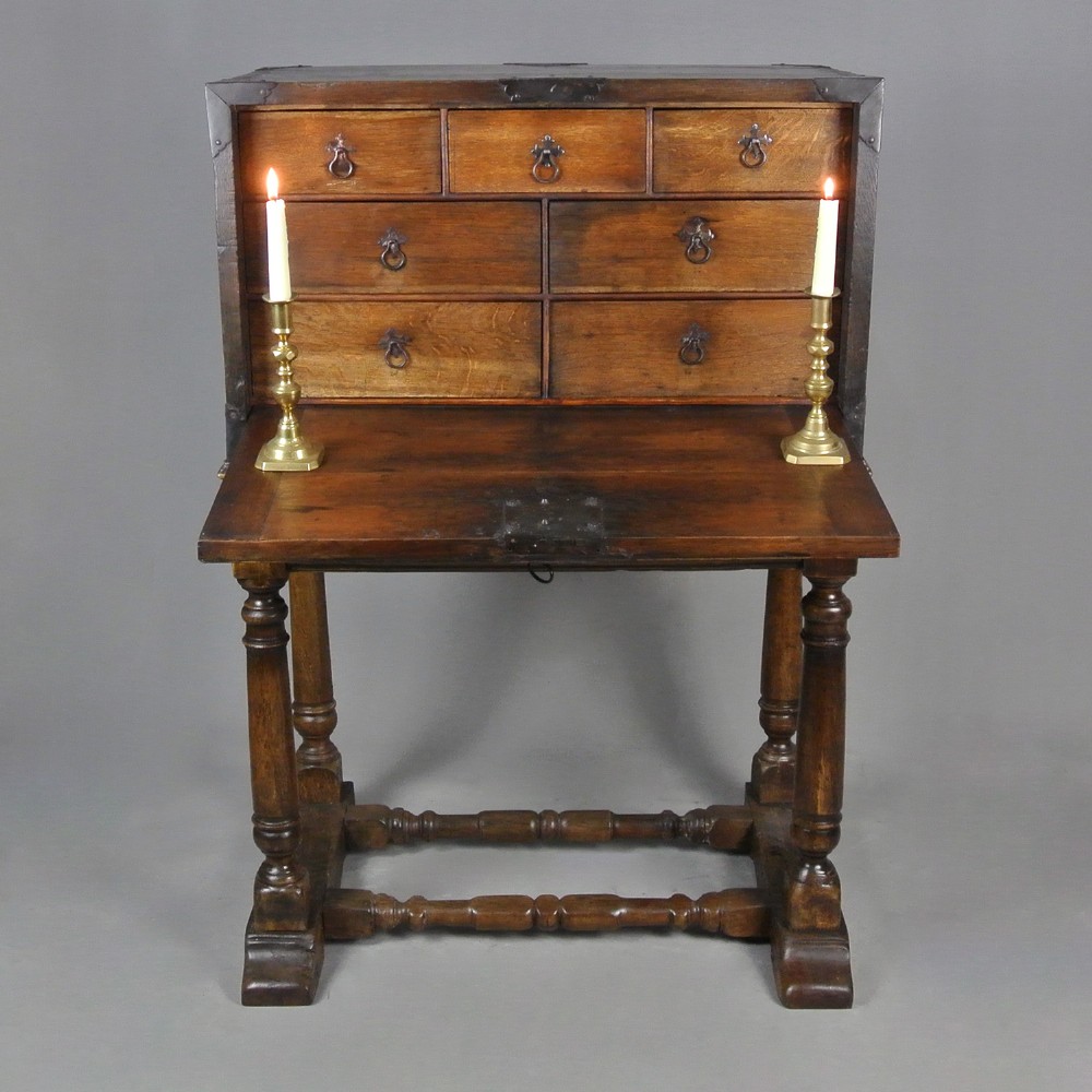 A 17th-century dark oak strong box featuring a fall-front desk opening to reveal small internal drawers, resting on a turned stand with stretchers.