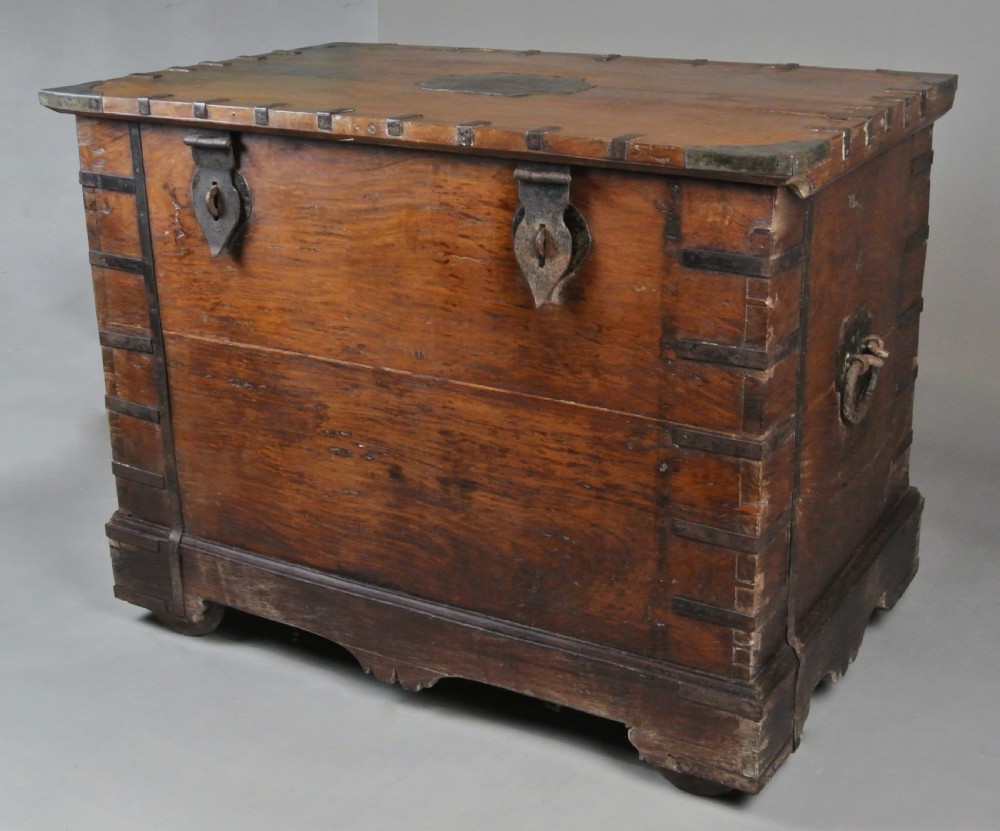 A large brown teak wood chest reinforced with dark iron straps, corner brackets, side handles, and front latches.