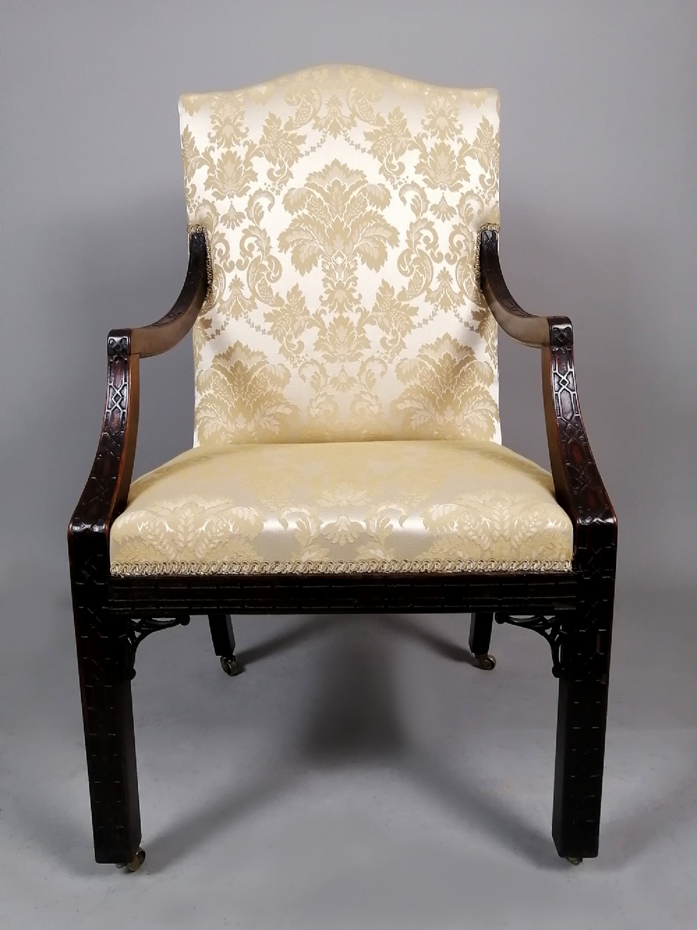 A George II mahogany library chair upholstered in cream damask fabric featuring carved Chinese Chippendale fretwork on the legs and arms.